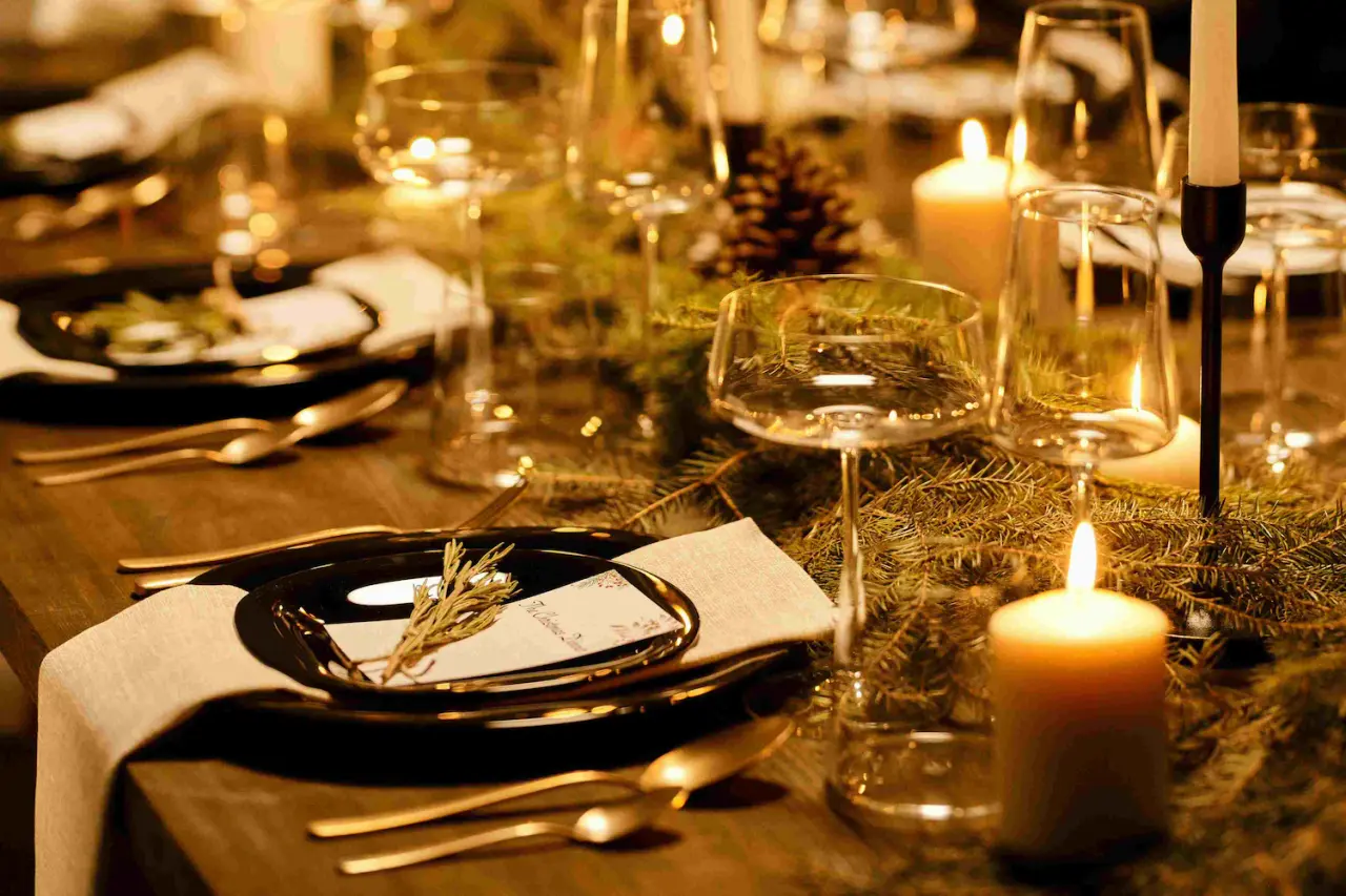 Mesa elegantemente decorada con vajilla negra, copas de cristal, velas encendidas y ramas de pino para una cena