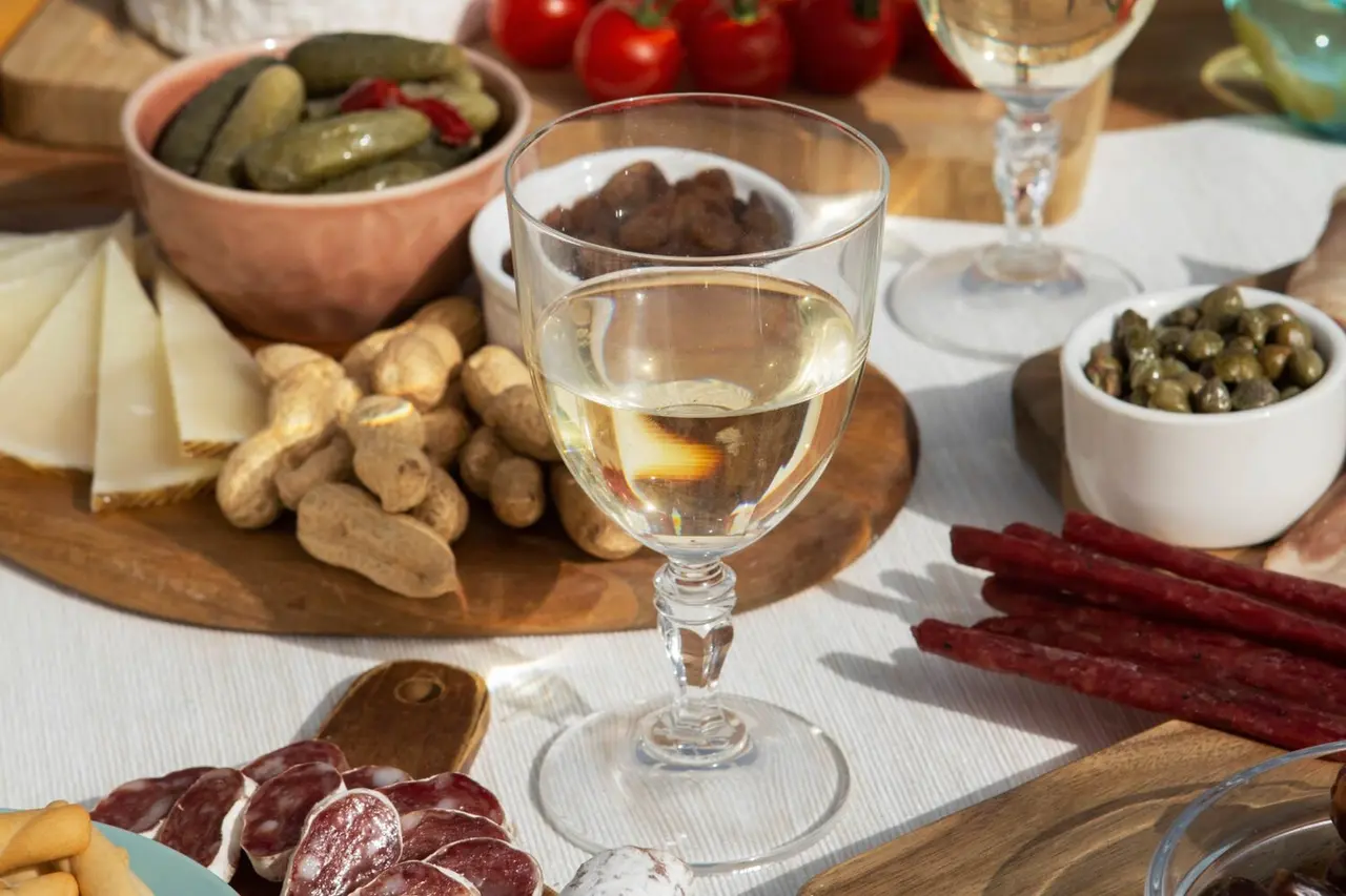 Mesa con copa de vino blanco, queso, embutidos, frutos secos, encurtidos y tomates cherry.