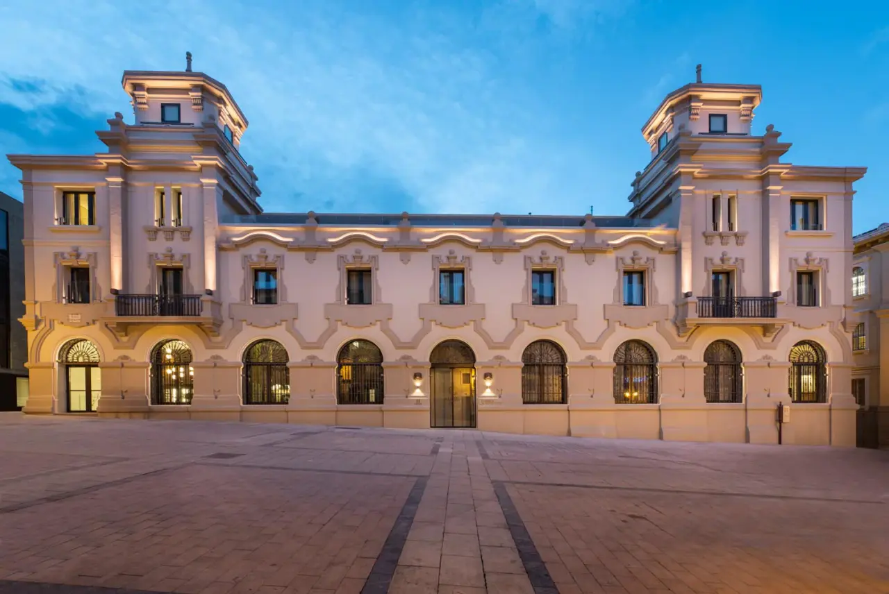 Fachada iluminada de un edificio histórico de estilo neoclásico con torres y arcos en una plaza al atardecer.