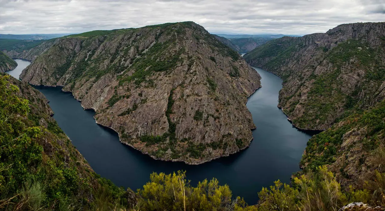 Vista panorámica de un río serpenteante rodeado de montañas rocosas y vegetación bajo un cielo nublado.