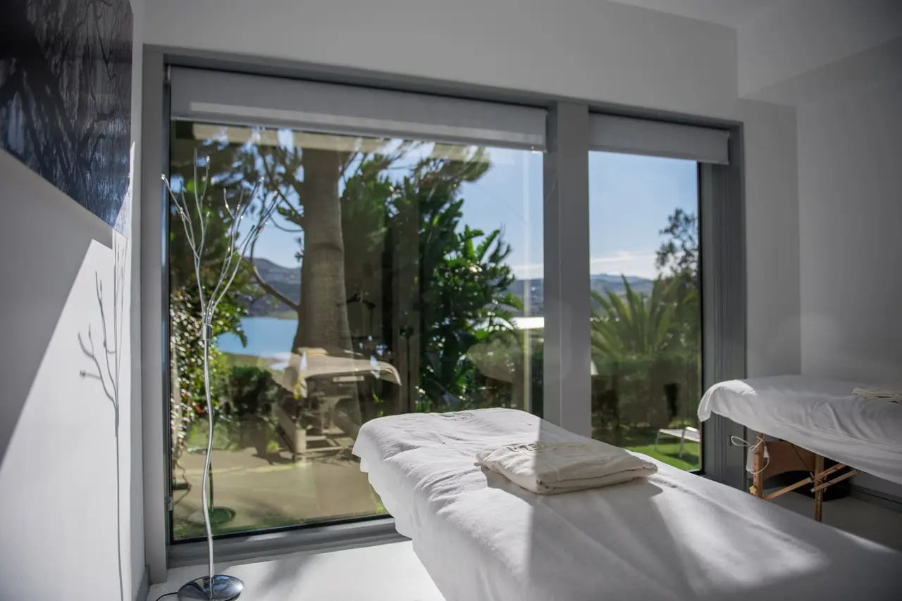 Habitación con dos camillas de masaje junto a una gran ventana con vistas al jardín y al mar.