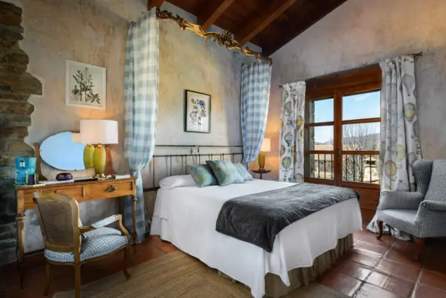 Habitación con cama doble, cabecero de forja, dosel con cortinas a cuadros, escritorio de madera con silla, butaca