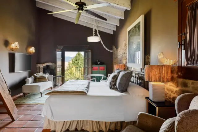 Vista de una habitación de hotel con cama grande vestida de blanco, vigas de madera en el techo, ventilador de techo