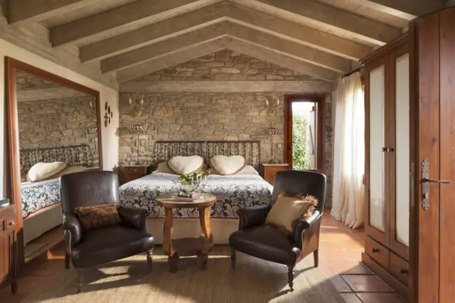 Habitación con techo abuhardillado de vigas de madera, paredes de piedra vista, cama doble con cabecero de forja