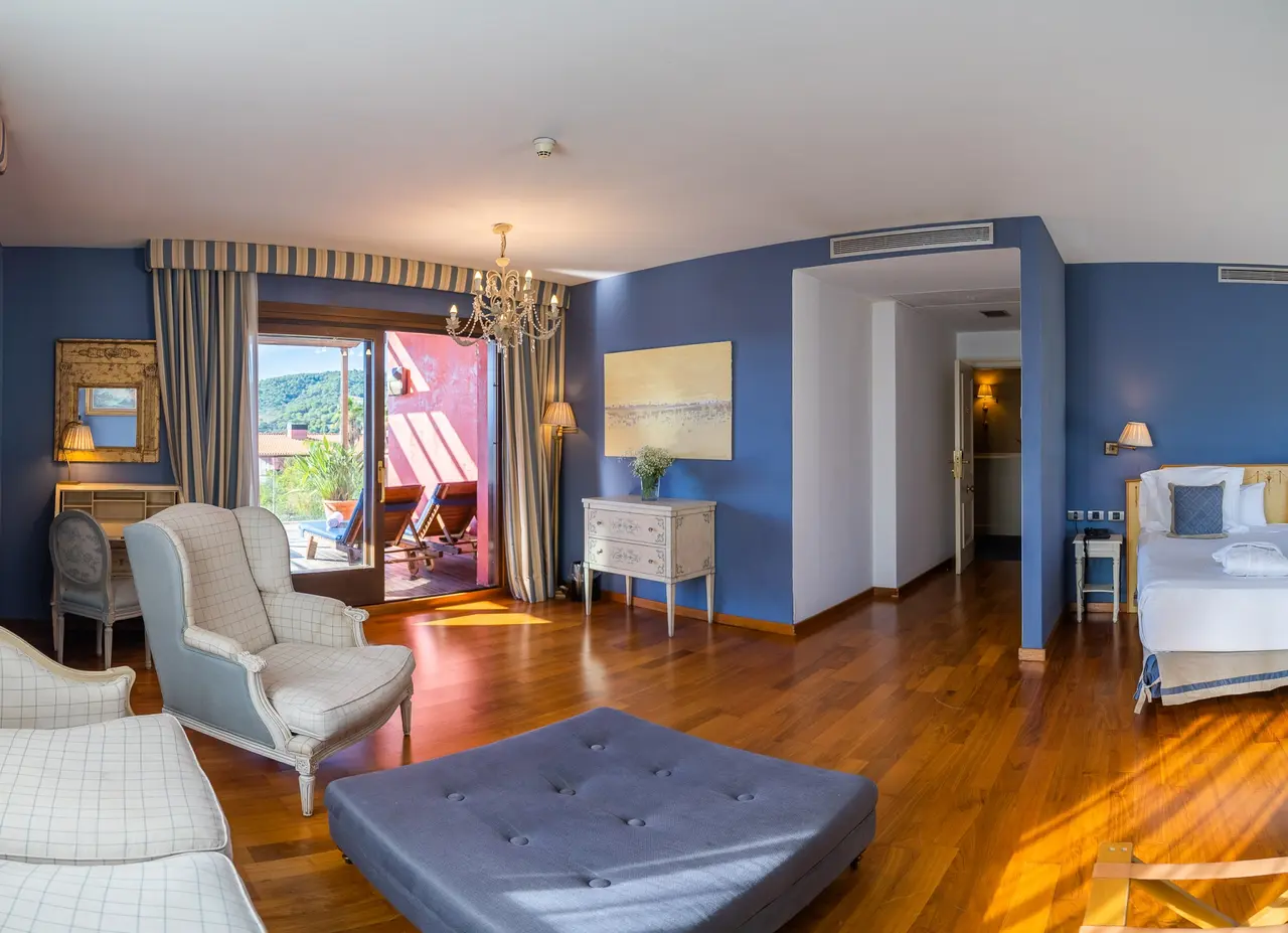 Interior de una suite con paredes azules, suelo de madera, zona de estar con sillones y otomana, cama a la derecha