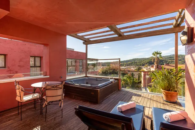 Jacuzzi rectangular empotrado en una terraza privada de madera con pérgola, rodeado de paredes de color terracota, mesa