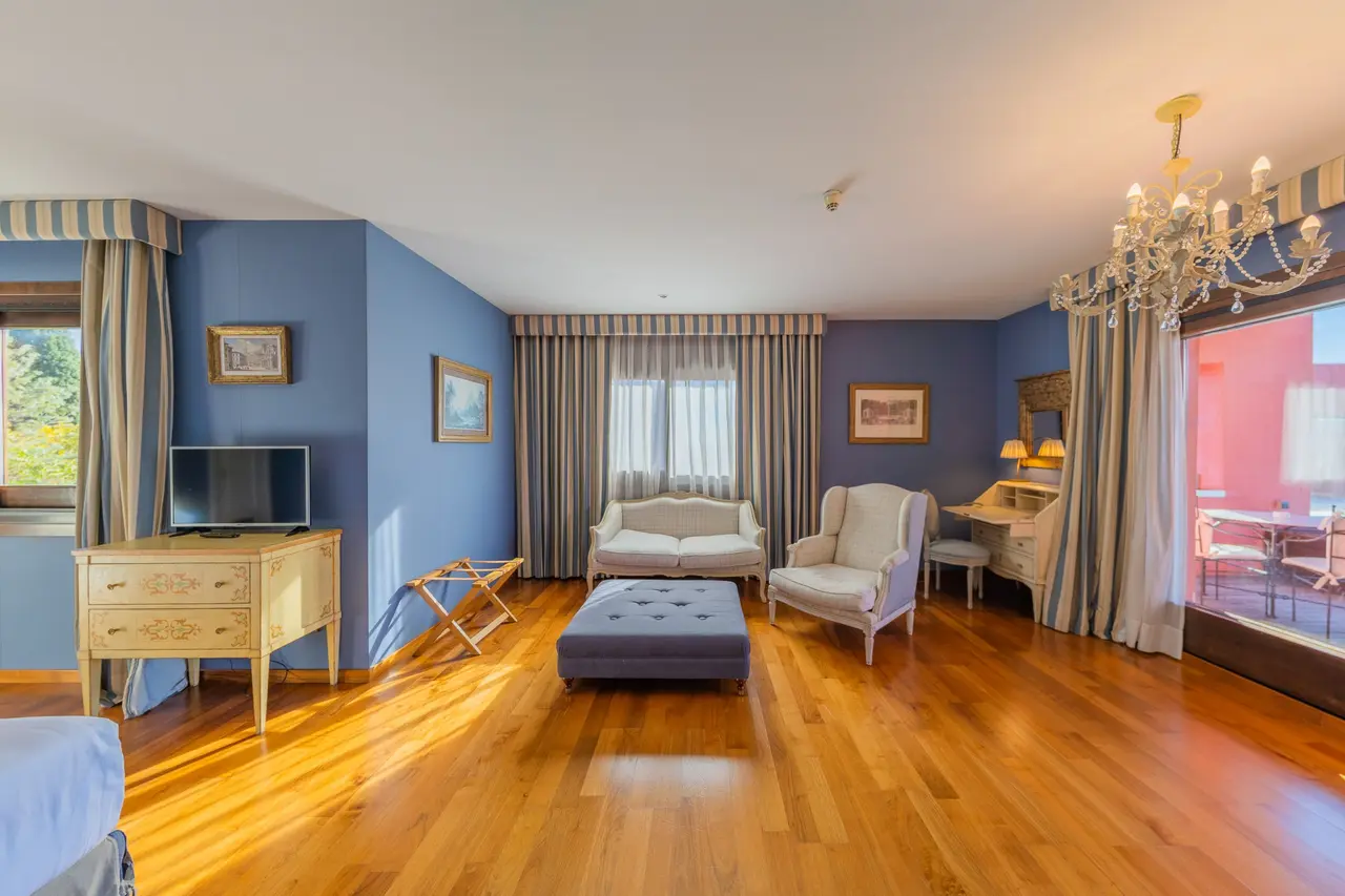 Vista de una suite de hotel con paredes azules, suelo de parquet, sofá, sillón, otomana, escritorio, televisor