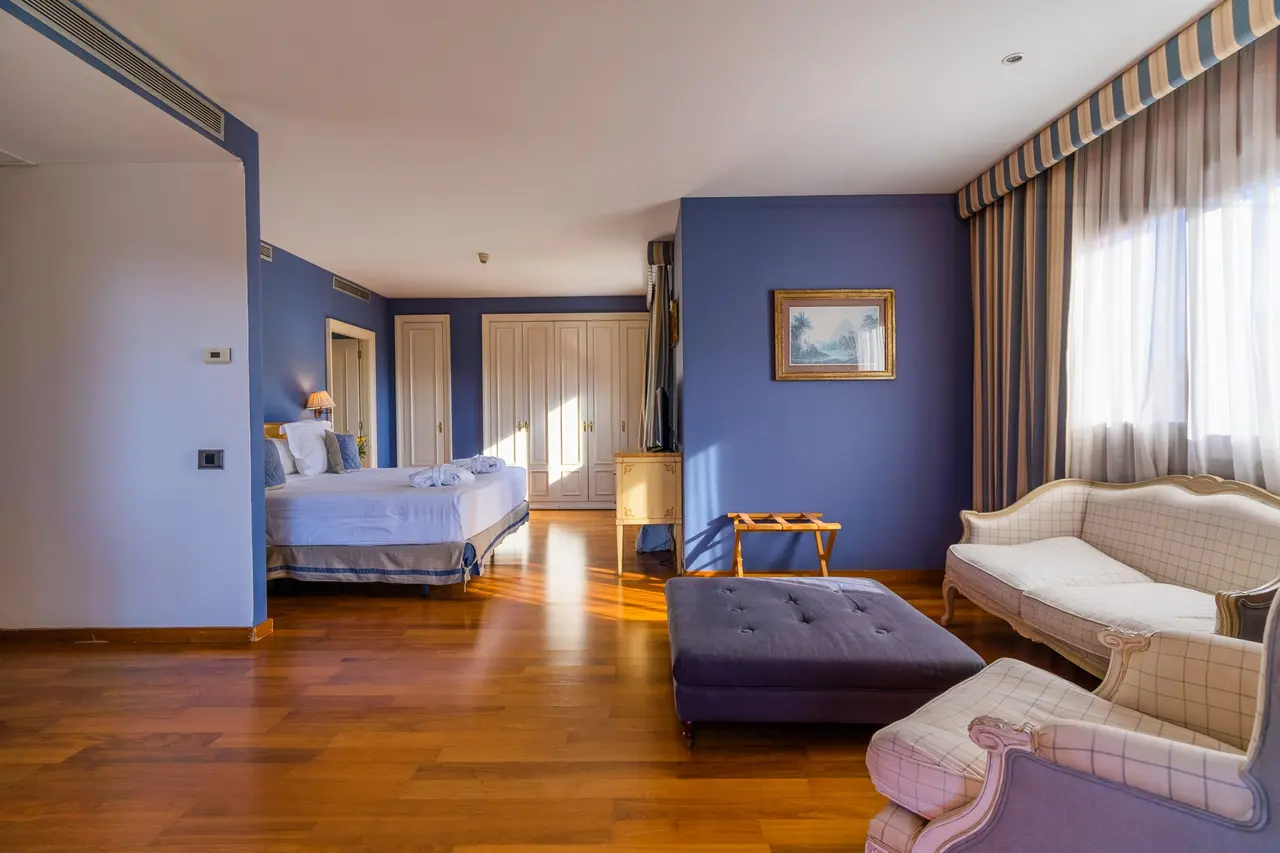 Vista de una suite de hotel con paredes azules, suelo de madera, una cama grande al fondo, un sofá y sillones