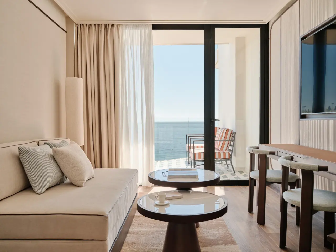 Interior de una junior suite con sofá beige, mesa de centro redonda, escritorio con sillas y gran puerta corredera