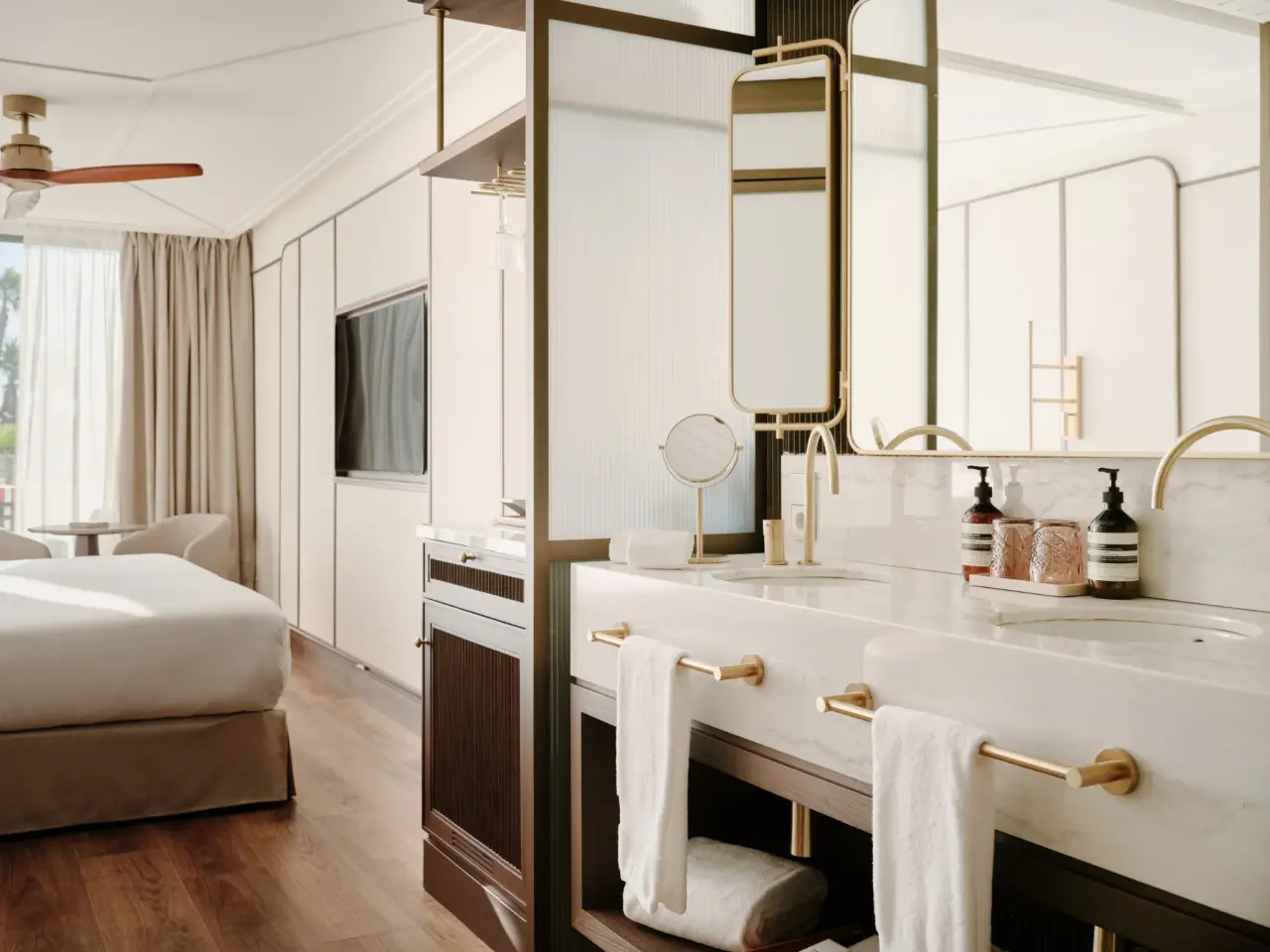 Vista de un baño moderno en suite con doble lavabo de mármol blanco, grifería dorada, toallas blancas y espejo grande.