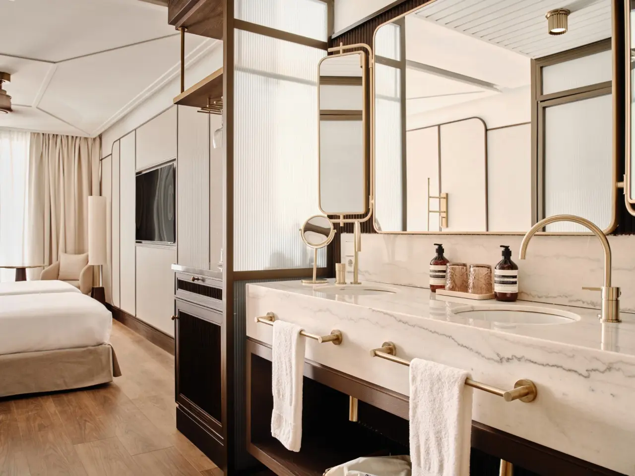 Baño de suite con doble lavabo de mármol blanco, grifería dorada, espejos grandes y toallas blancas.
