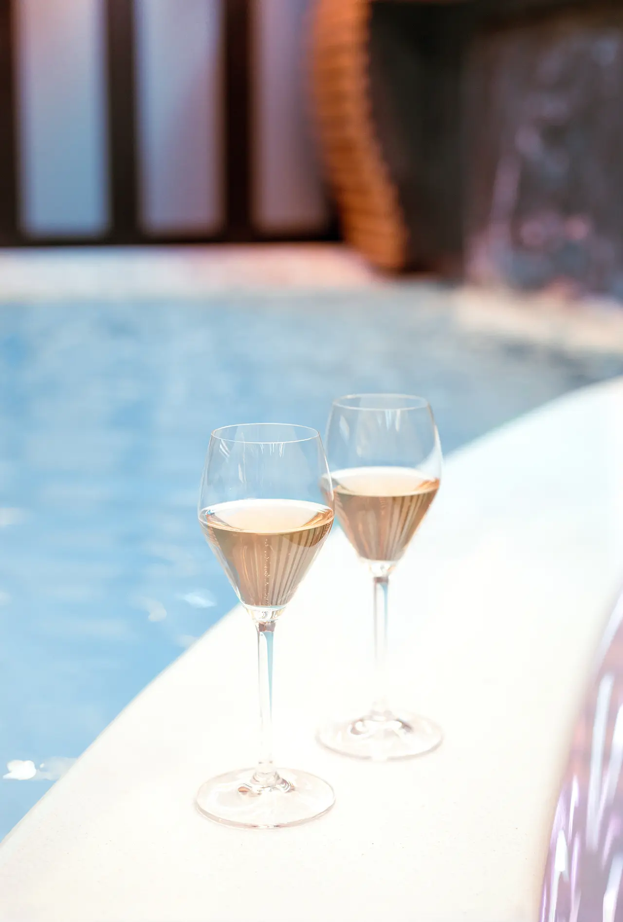 Dos copas de vino rosado sobre el borde de una piscina con agua azul.