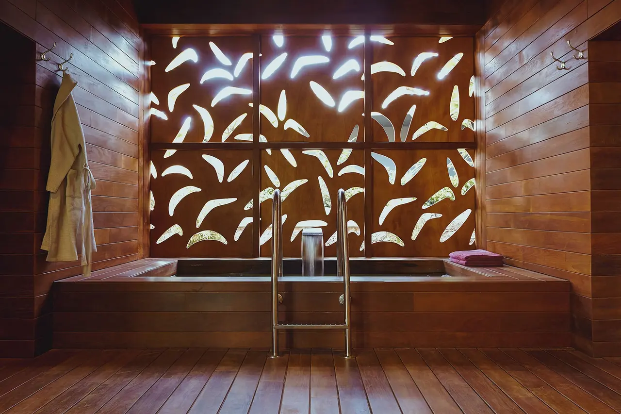 Jacuzzi interior de madera con escalera metálica, pared decorativa calada iluminada y albornoz colgado en la pared.