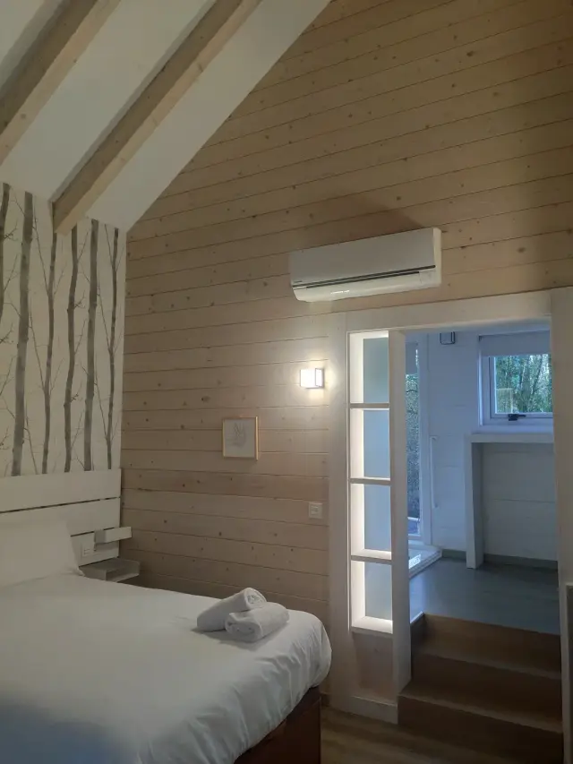 Interior de una habitación con paredes y techo de madera clara, cama con sábanas blancas y toallas enrolladas, papel