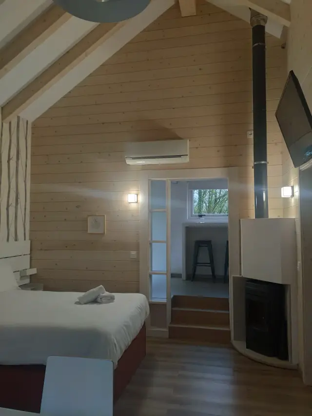 Interior de una habitación con paredes y techo de madera clara, cama doble con sábanas blancas, chimenea con estufa