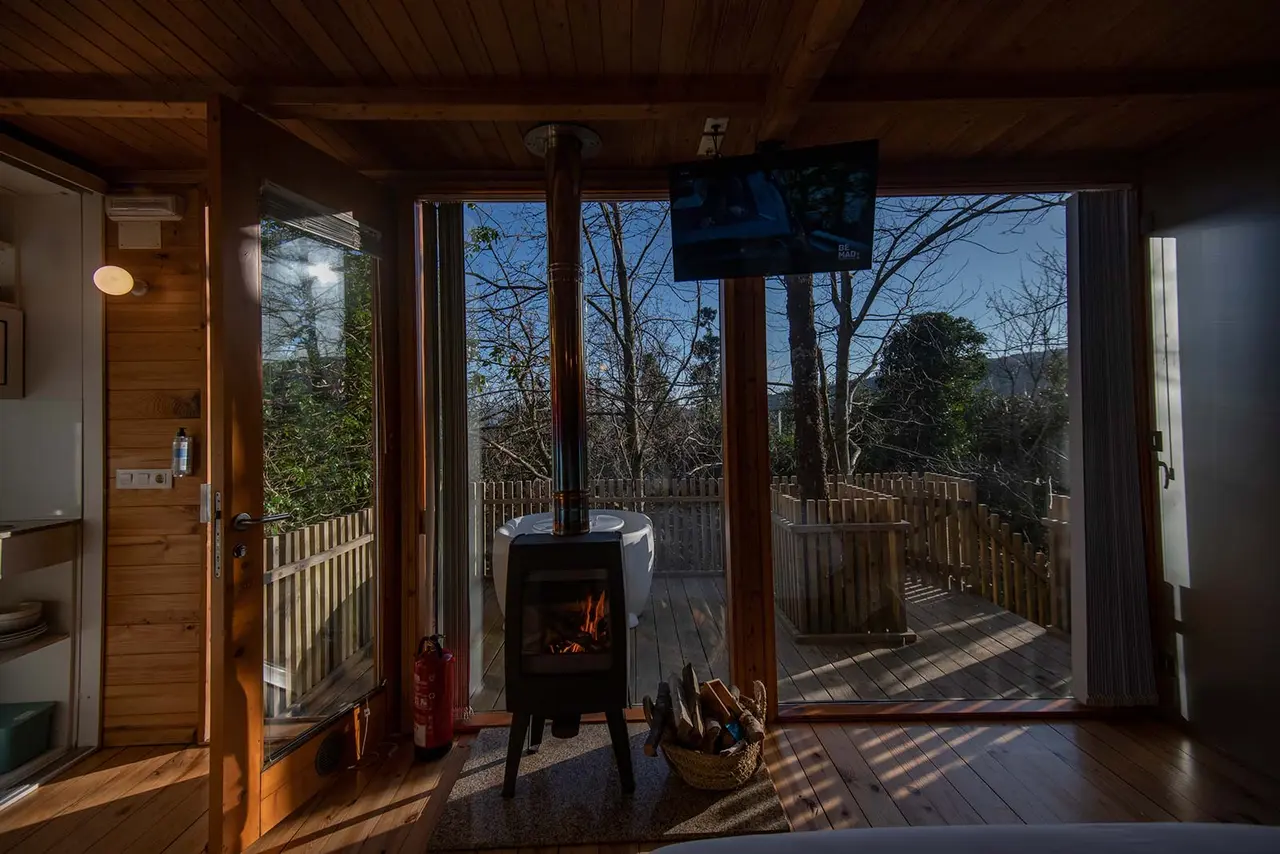 Vista interior de una cabaña de madera con chimenea encendida, suelo de madera y puertas abiertas que dan