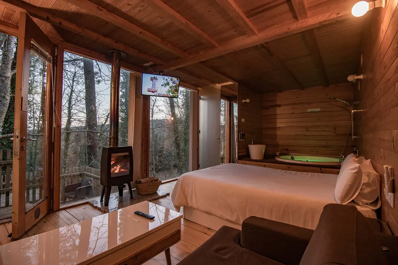 Interior de una cabaña de madera con cama grande, chimenea encendida, sofá, mesa de centro y jacuzzi visible