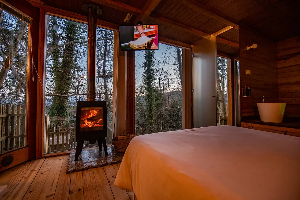 Interior de una cabaña de madera con cama grande en primer plano, chimenea encendida, suelo de madera, grandes