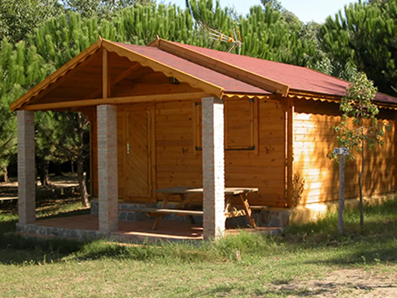 Vista exterior de un bungalow de madera con techo rojo y porche cubierto sostenido por pilares de ladrillo.