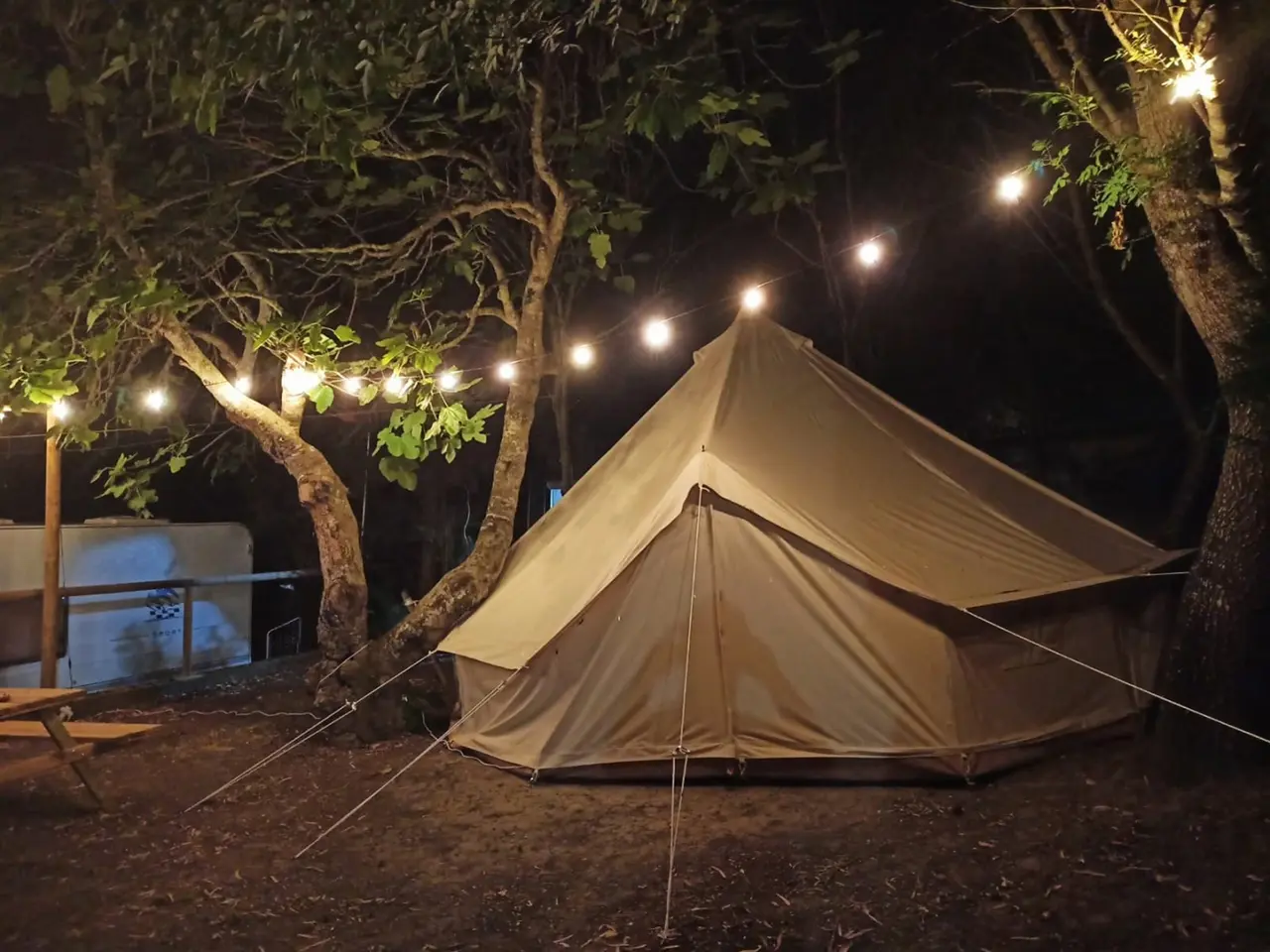 Vista nocturna de una tienda de campaña tipo glamping de color beige, iluminada por una guirnalda de luces colgadas