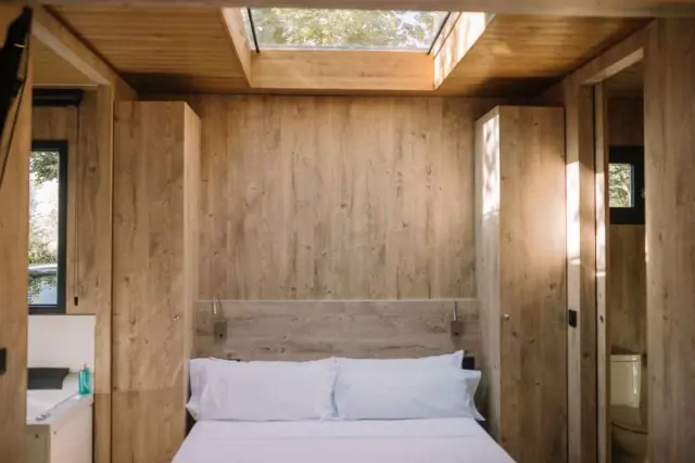 Interior de un bungalow con paredes y techo de madera clara, una cama doble con sábanas y almohadas blancas, armarios