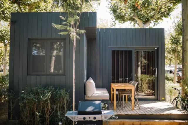 Vista exterior de un bungalow moderno de color gris oscuro con terraza de madera, mesa y sillas, barbacoa y vegetación