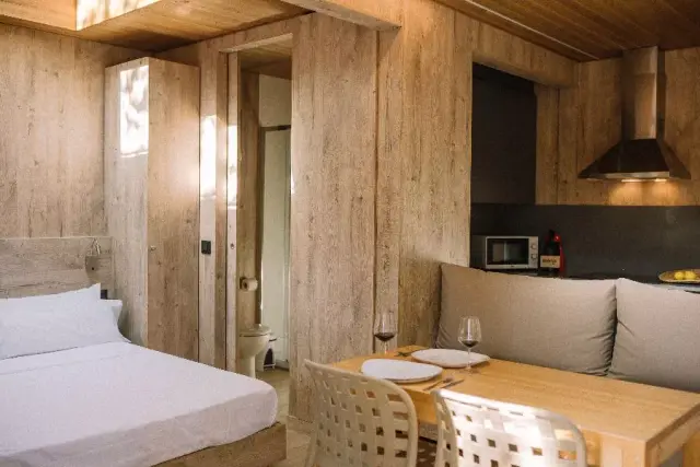 Interior de un bungalow con paredes y techo de madera clara. A la izquierda se ve una cama doble con sábanas blancas