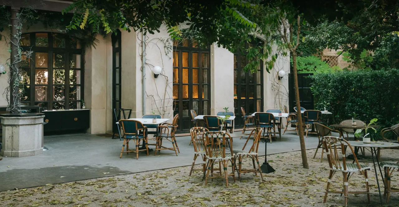 Patio exterior con mesas y sillas de madera dispuestas sobre suelo de grava, rodeado de vegetación y fachada