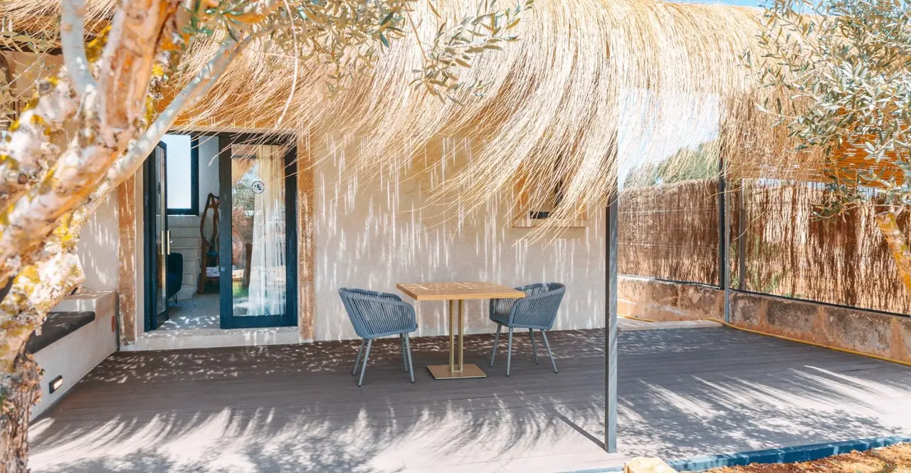 Terraza exterior de una suite con suelo de madera, mesa redonda con dos sillas, techo de paja y vistas a un olivo