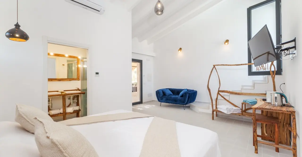 Interior de una suite con cama grande en primer plano, sofá azul, mesa de madera rústica con hervidor, televisión