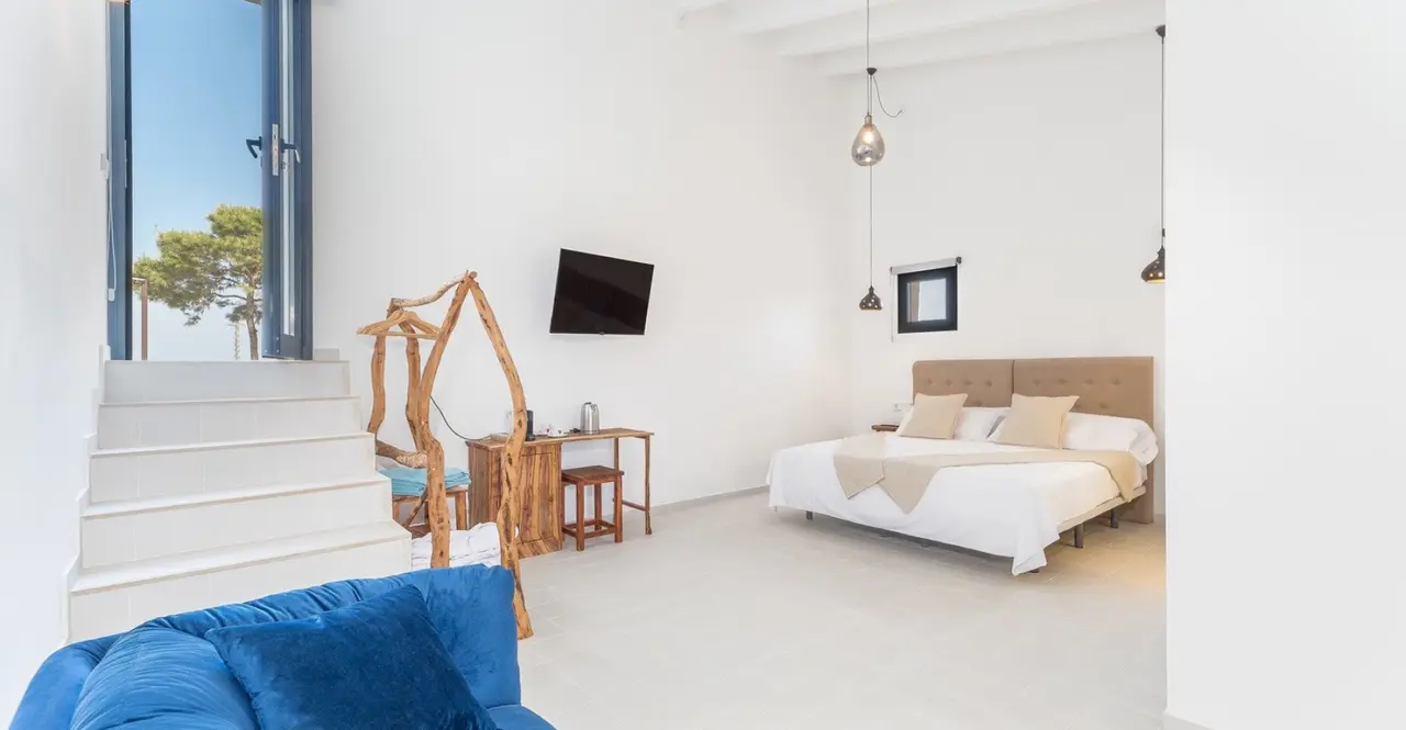 Interior de una suite con paredes blancas, suelo claro, cama doble con cabecero de madera y ropa de cama blanca