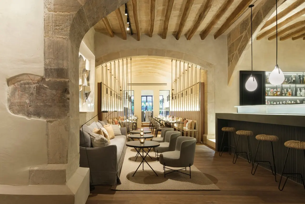 Salón de hotel con sofás, sillones, mesas y barra de bar bajo arcos de piedra y techo de vigas de madera.