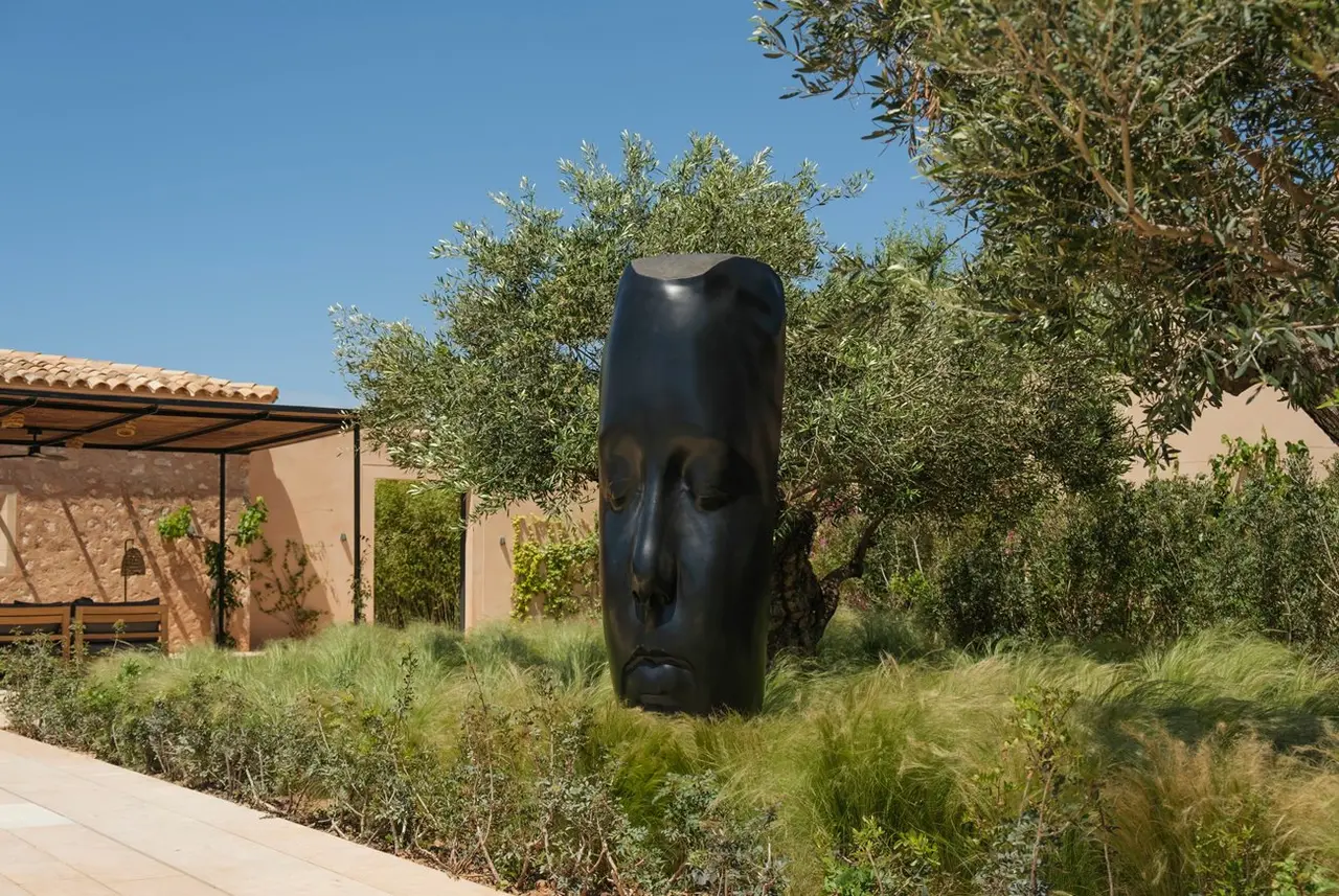 Escultura negra de una cabeza en un jardín exterior con vegetación, árboles y un edificio al fondo bajo un cielo azul.