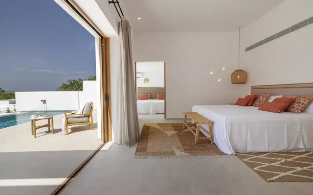 Habitación rural con cama grande vestida de blanco y cojines terracota, suelo de baldosas claras, alfombras de yute