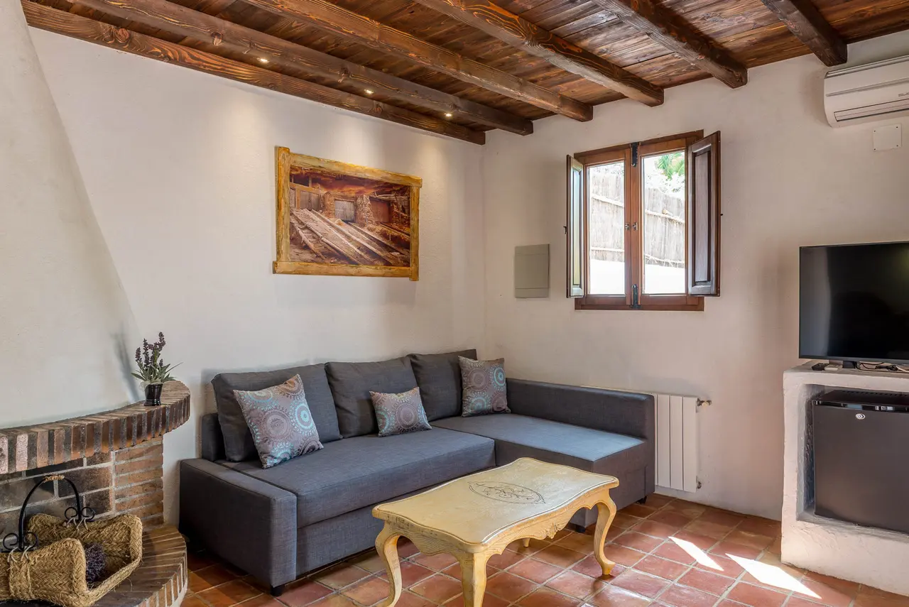 Salón rústico con sofá gris en L, mesa de centro, chimenea de ladrillo, ventana y vigas de madera en el techo.