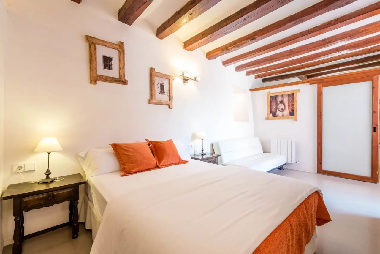 Habitación doble con cama grande, paredes blancas, vigas de madera en el techo y muebles de madera oscura.