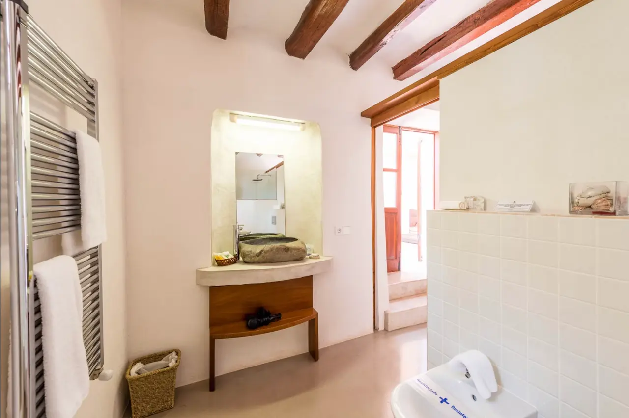 Baño con lavabo de piedra sobre mueble de madera, espejo iluminado, toallero eléctrico, inodoro y vigas de madera