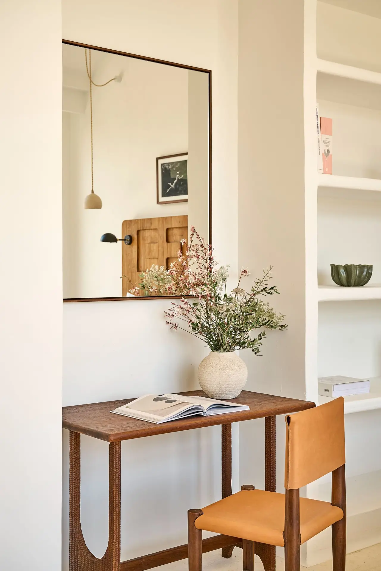 Interior de una habitación con una consola de madera, silla de cuero marrón, jarrón con flores secas y un gran espejo