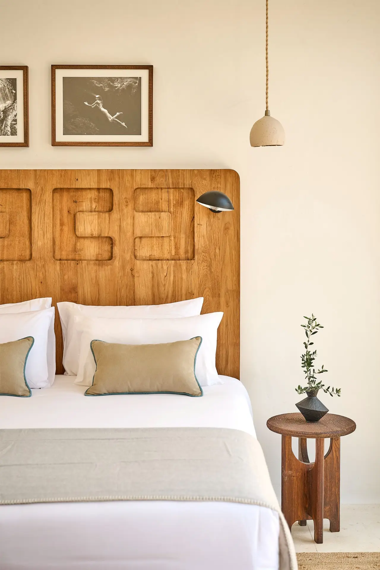 Habitación con cama grande vestida de blanco, cabecero de madera tallada, dos cuadros enmarcados en la pared, mesita