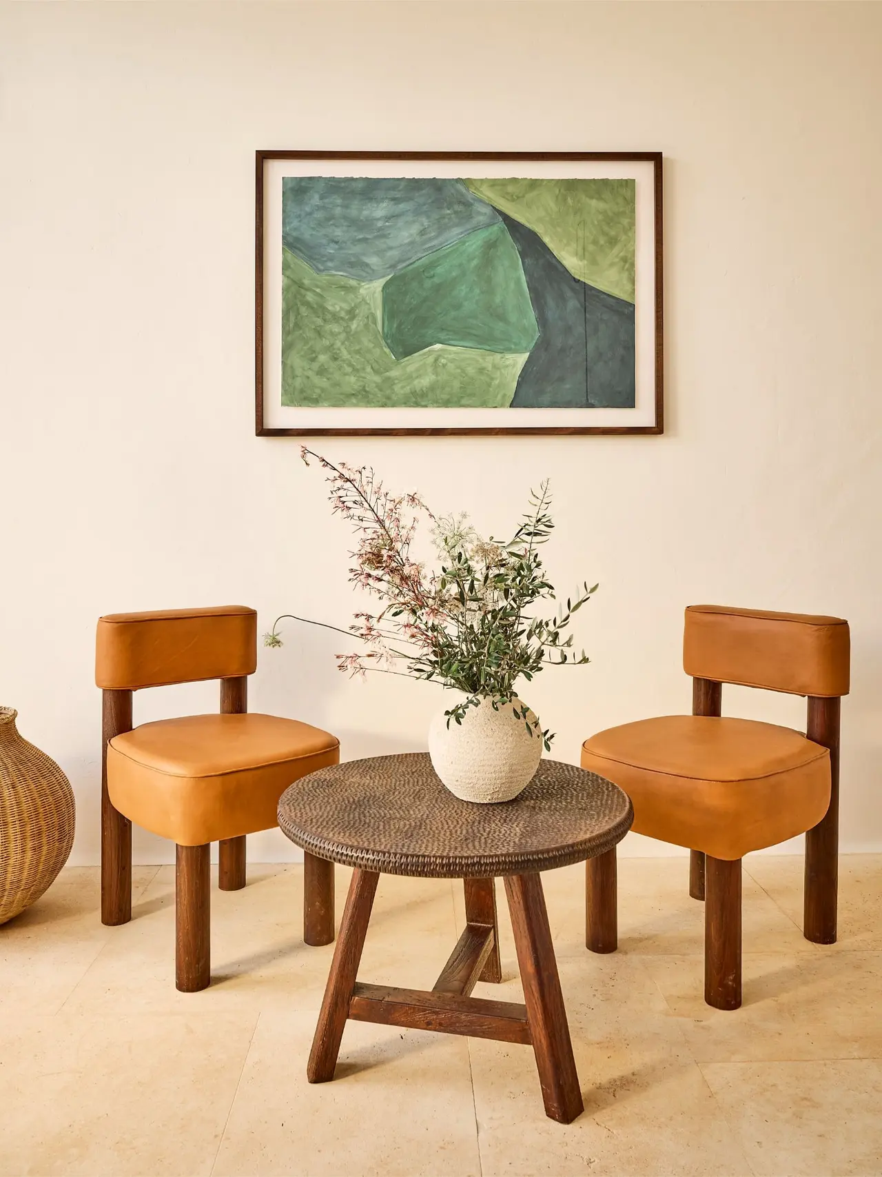 Interior de una habitación con dos sillas de cuero marrón, una mesa redonda de madera con un jarrón de flores secas