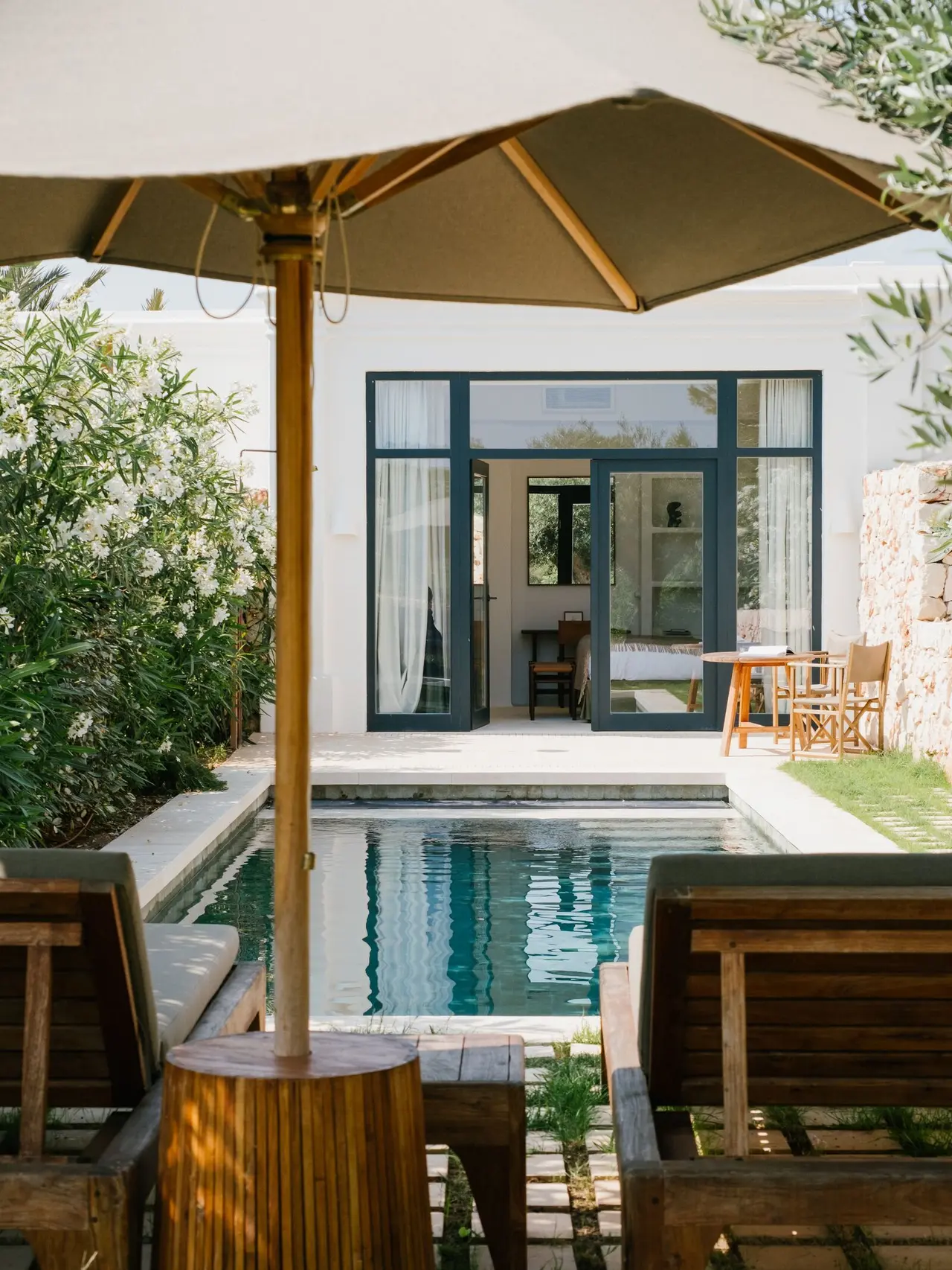 Piscina privada rectangular con agua turquesa, rodeada de tumbonas de madera, una mesa auxiliar y una gran sombrilla.