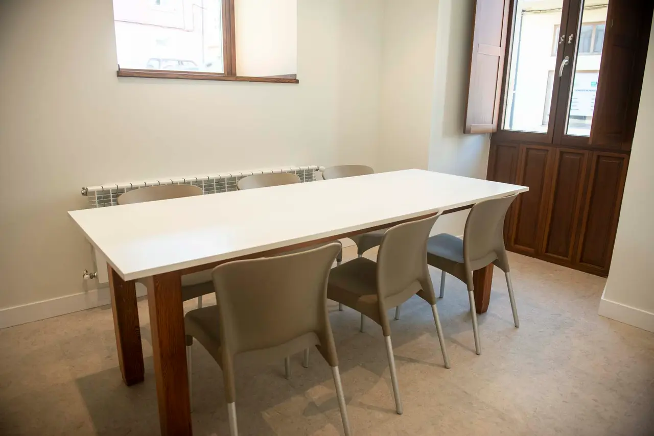 Habitación interior con una mesa rectangular blanca grande rodeada de seis sillas beige, suelo de baldosas claras