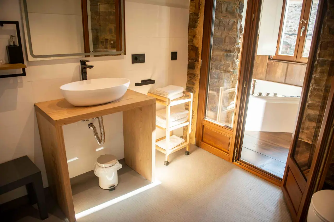 Baño con lavabo de cerámica blanca sobre mueble de madera, espejo grande, toallero con toallas y puerta abierta que da