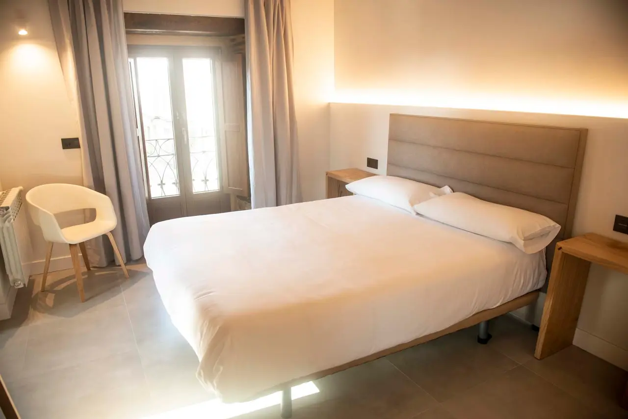 Vista de una suite con cama doble grande vestida de blanco, cabecero acolchado, mesitas de noche de madera, silla