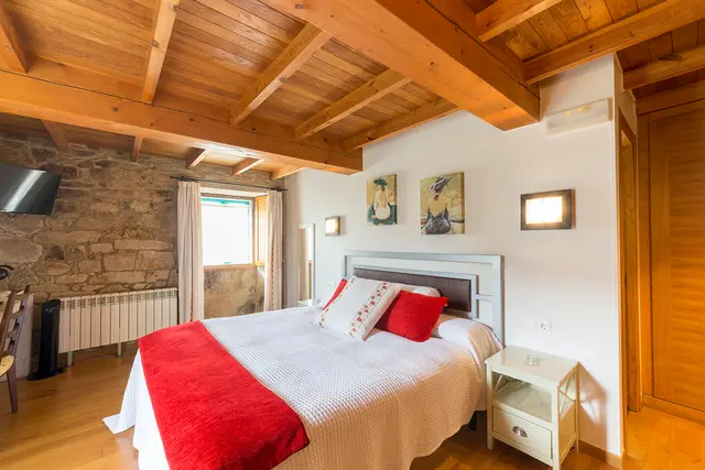 Habitación con cama doble vestida con colcha blanca y manta roja, cabecero gris, vigas de madera en el techo, pared de p