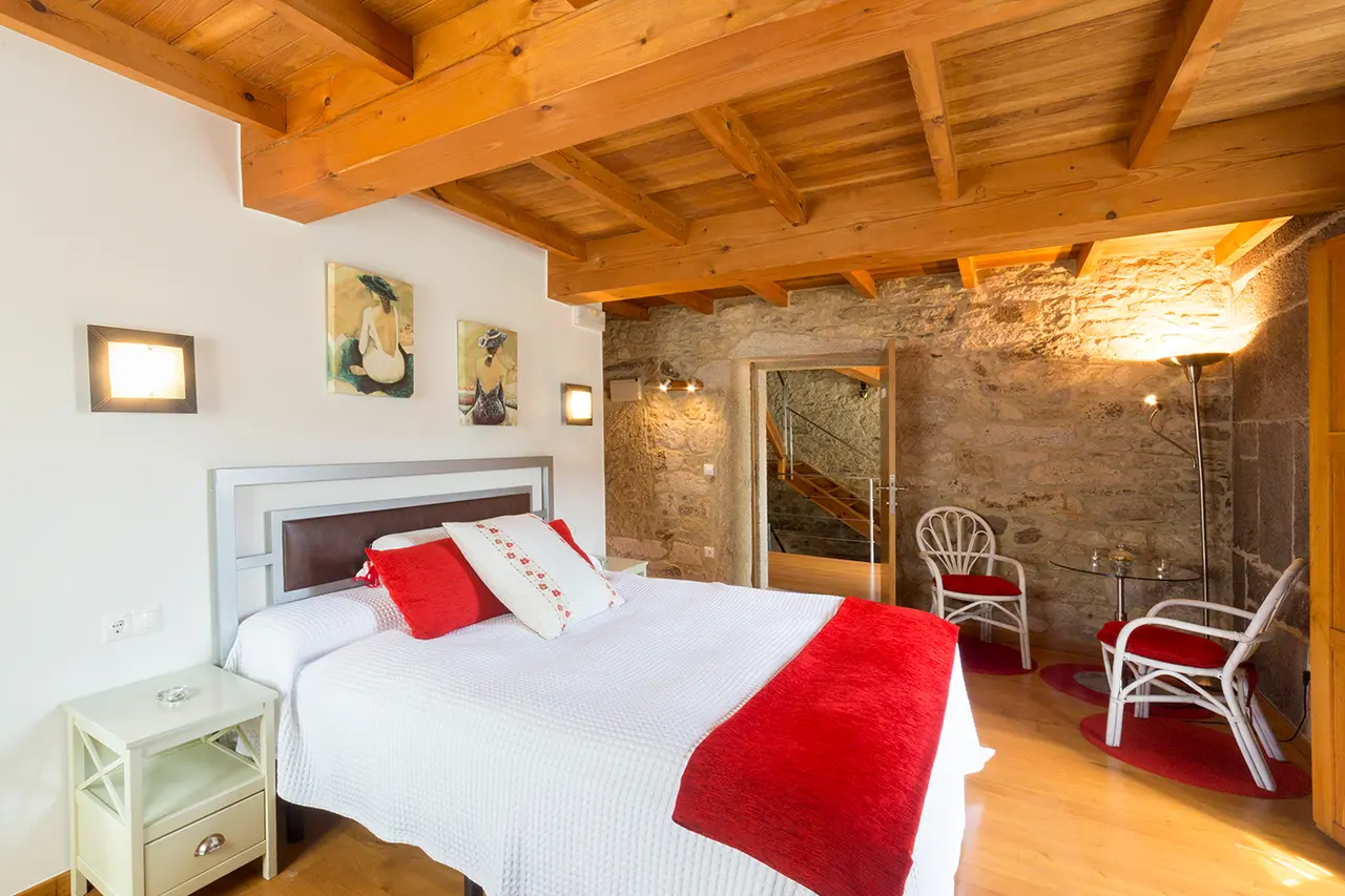 Habitación con cama doble vestida con colcha blanca y manta roja, cabecero plateado, mesitas de noche, sillas blancas