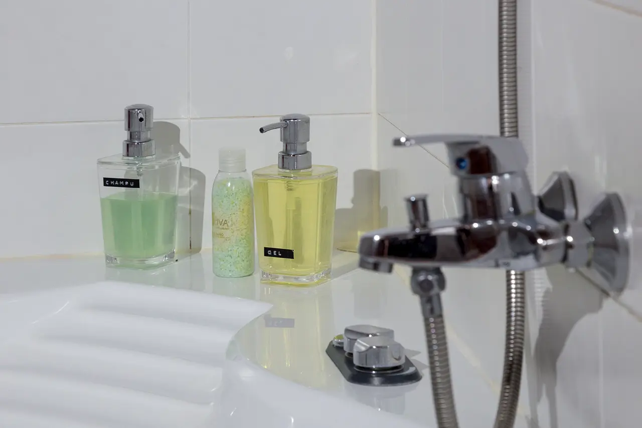 Detalle de una bañera blanca con grifo cromado y varios dispensadores de gel, champú y sales de baño sobre el borde.