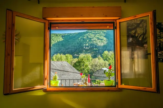 Vista a través de una ventana de madera abierta con persianas enrollables, mostrando un paisaje de montañas verdes