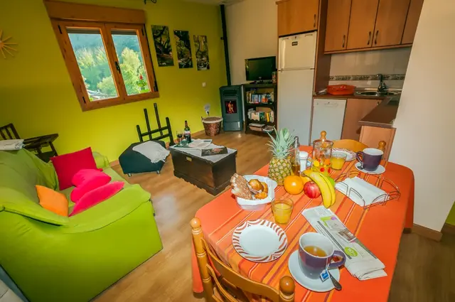 Interior de una habitación con sala de estar y cocina. Se ve un sofá verde con cojines de colores, una mesa de comedor