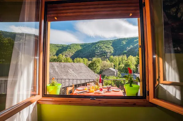 Vista desde una ventana abierta hacia un paisaje de montañas verdes y un pueblo con tejados, con una mesa preparada